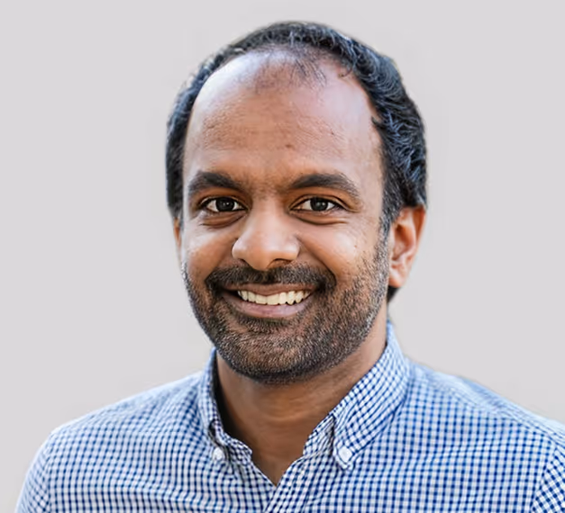 A smiling man with dark hair and a checked shirt, standing in front of a neutral gray background