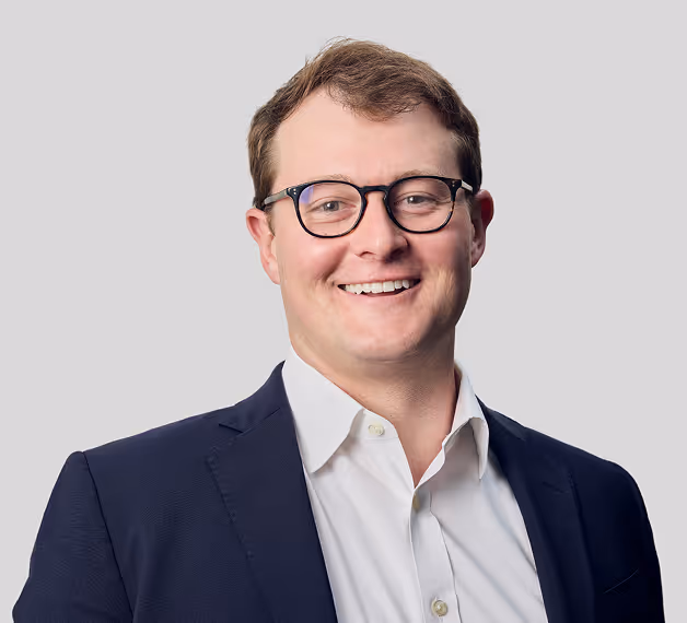 Professional headshot of smiling man with dark-framed glasses wearing navy blazer over white shirt