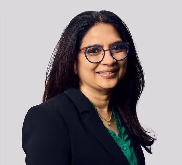 Professional headshot of woman with long dark hair wearing blue-framed glasses, green blouse, and black blazer