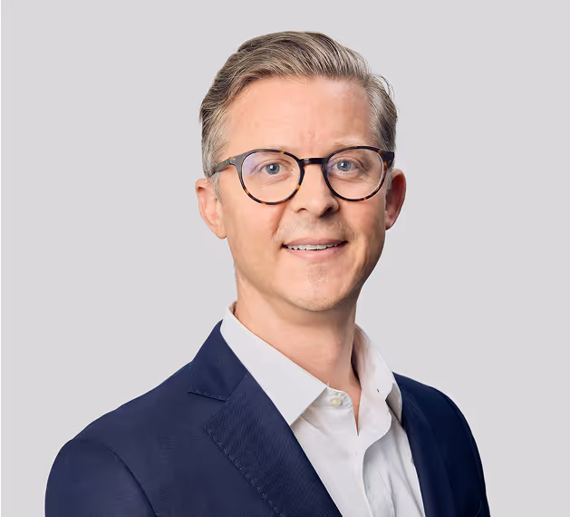 A professional headshot of a man in glasses wearing a navy blazer and white shirt, set against a light gray background