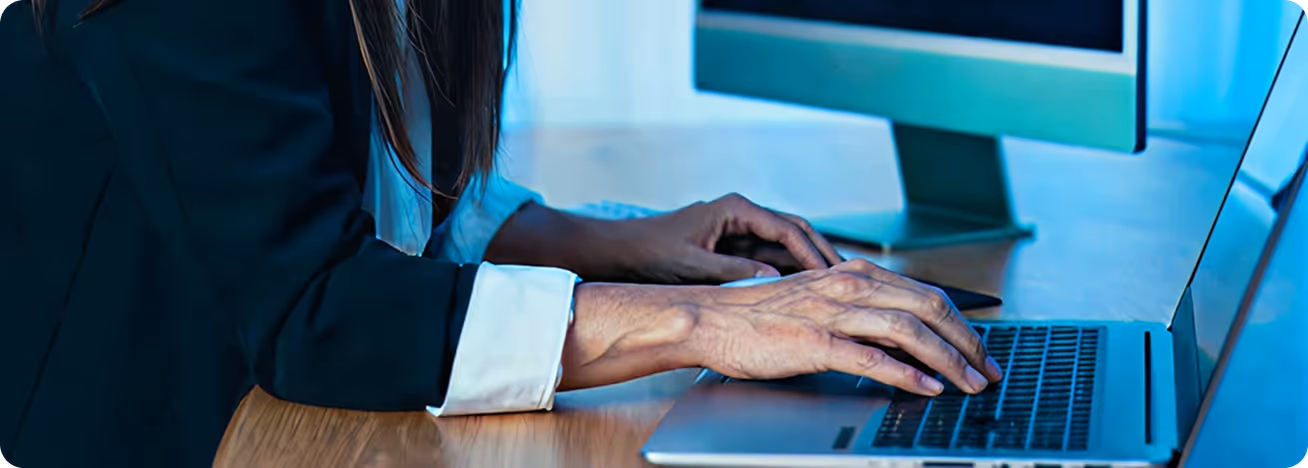 Close-up of two hands typing on laptops at a desk, one hand on a computer mouse and the other on a laptop keyboard