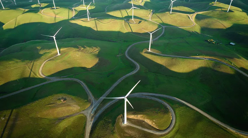 Aerial view of a green hilly landscape with multiple white wind turbines casting long shadows.