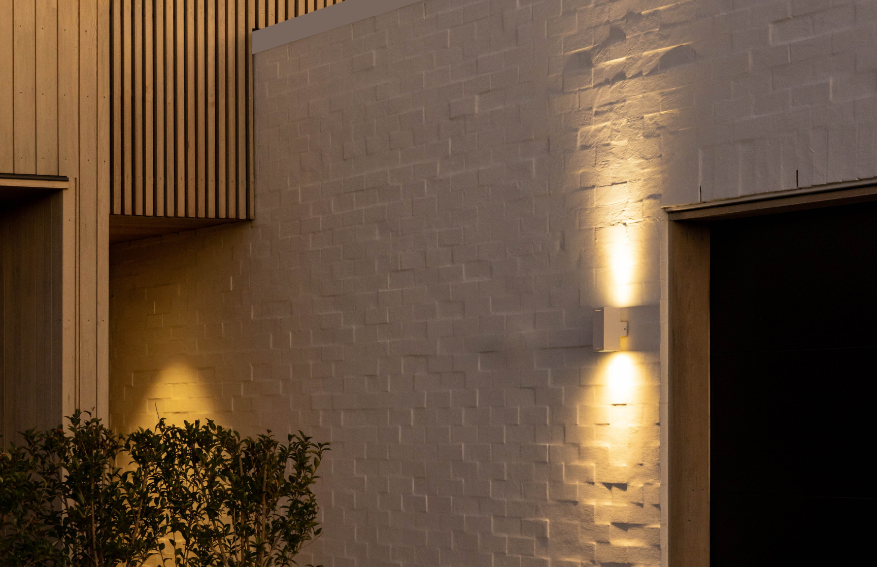 Exterior wall of a modern building with textured white bricks, vertical wooden paneling, warm wall lights, and shrubbery in the foreground.