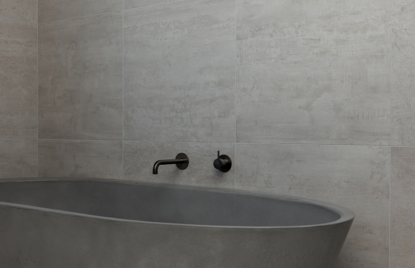 Modern matte gray freestanding bathtub against large gray tiled wall with black wall-mounted faucet and handle.