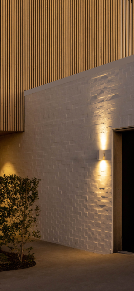 Evening view of a modern building corner with vertical wooden slats above a white brick wall illuminated by wall-mounted up and down lights, with a small tree nearby.