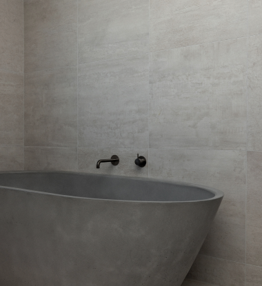 Minimalist gray stone bathtub with matte black wall-mounted faucet against beige tiled wall.