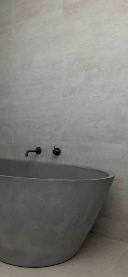 Modern bathroom with a matte gray freestanding bathtub against large beige tiled walls and dark wall-mounted faucet.