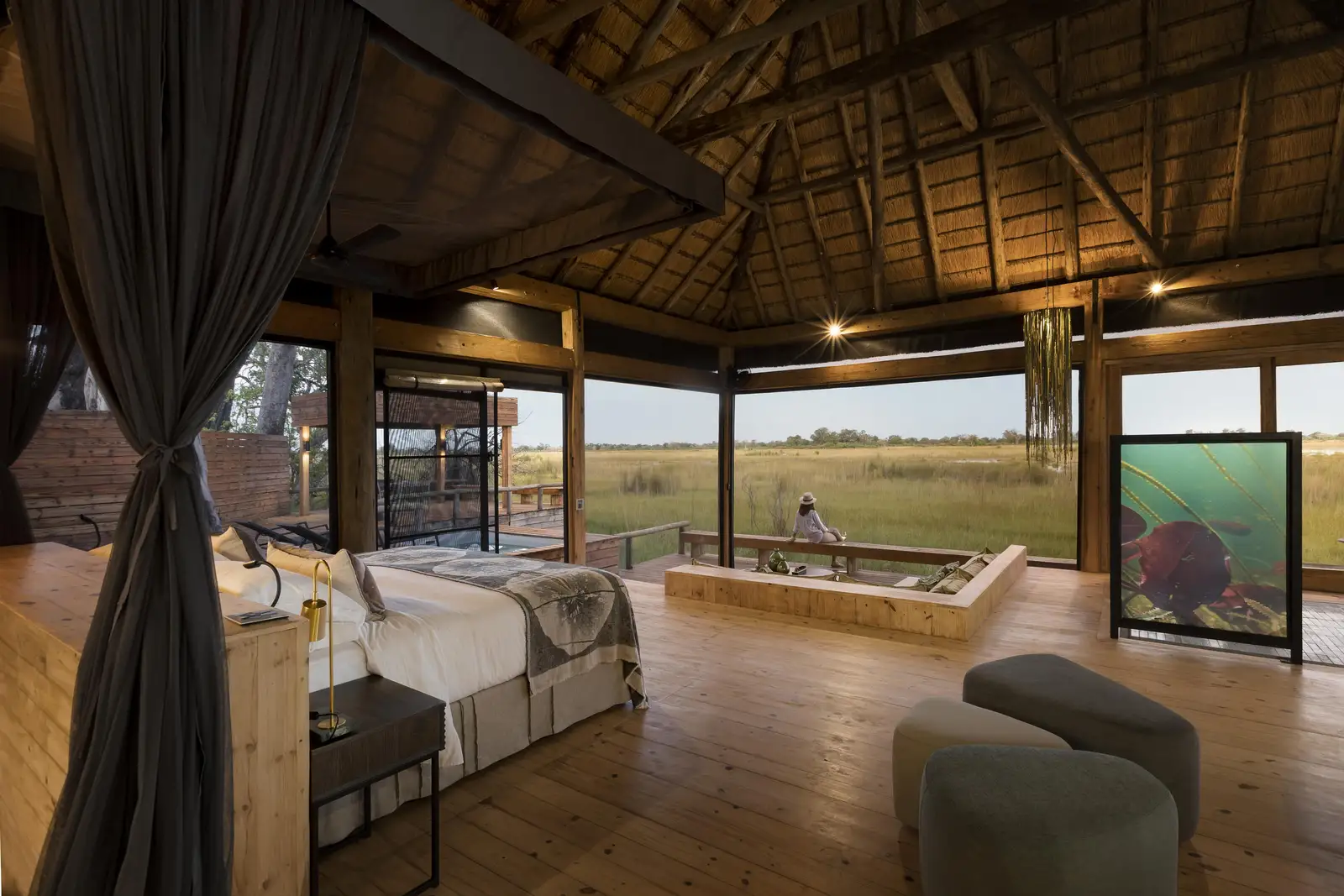 A luxury thatched-roof safari suite with expansive floor-to-ceiling views of an open grassy plain.