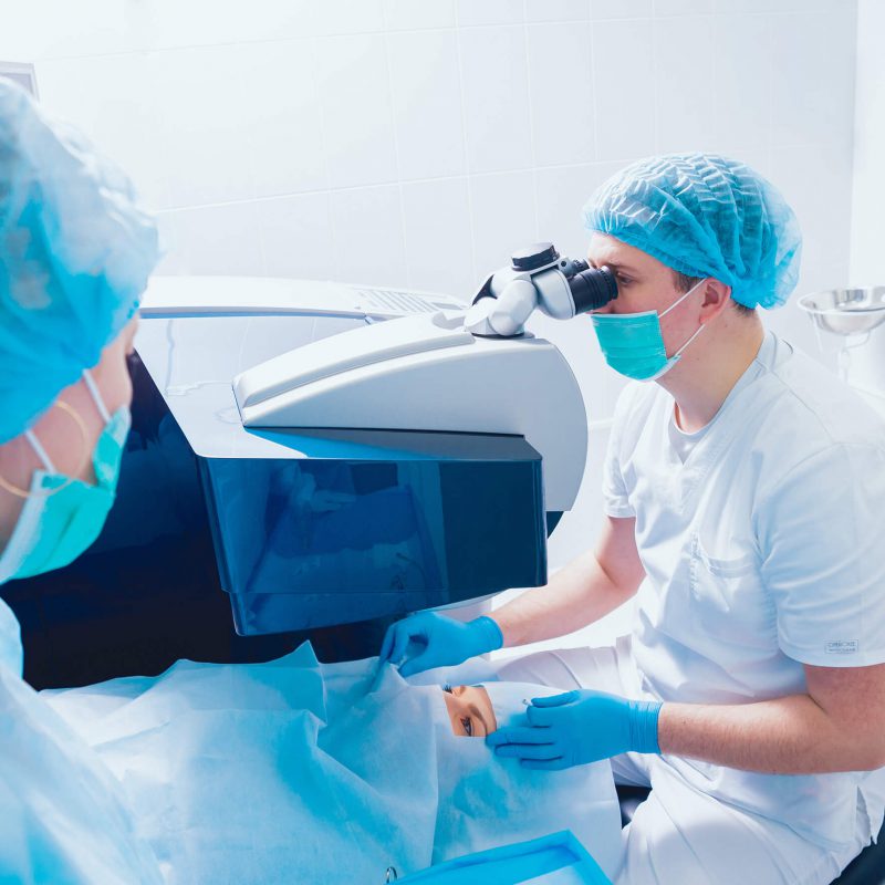 Surgeon wearing surgical scrubs and mask looking through a microscope while performing eye surgery on a patient.