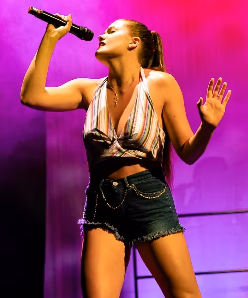 A performer in a striped top and denim shorts dances on a pink-lit stage.