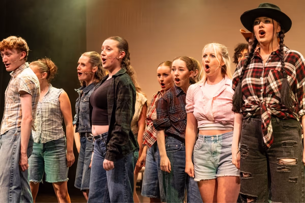 Performers in denim and checked shirts sing together on stage in an ensemble scene.