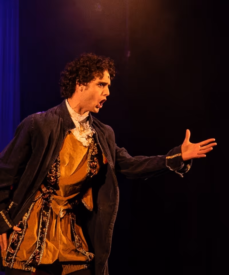 A performer in a period costume and velvet coat delivers a dramatic moment on stage under warm light.
