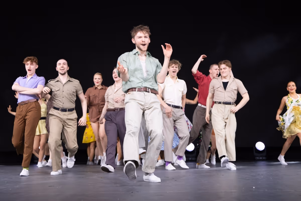 A company of performers in khaki costumes dances across the stage, led by a performer in a green shirt.