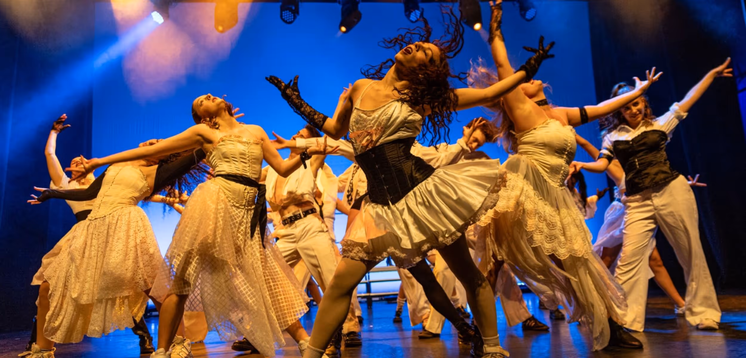 Dancers in lace dresses and corsets perform an energetic group number under blue stage light.