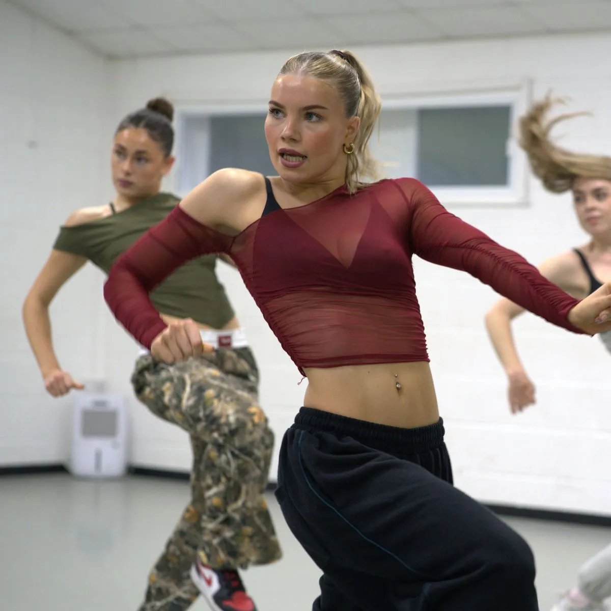 A dancer in a red mesh top leads a commercial dance routine in a studio, with two dancers behind her.