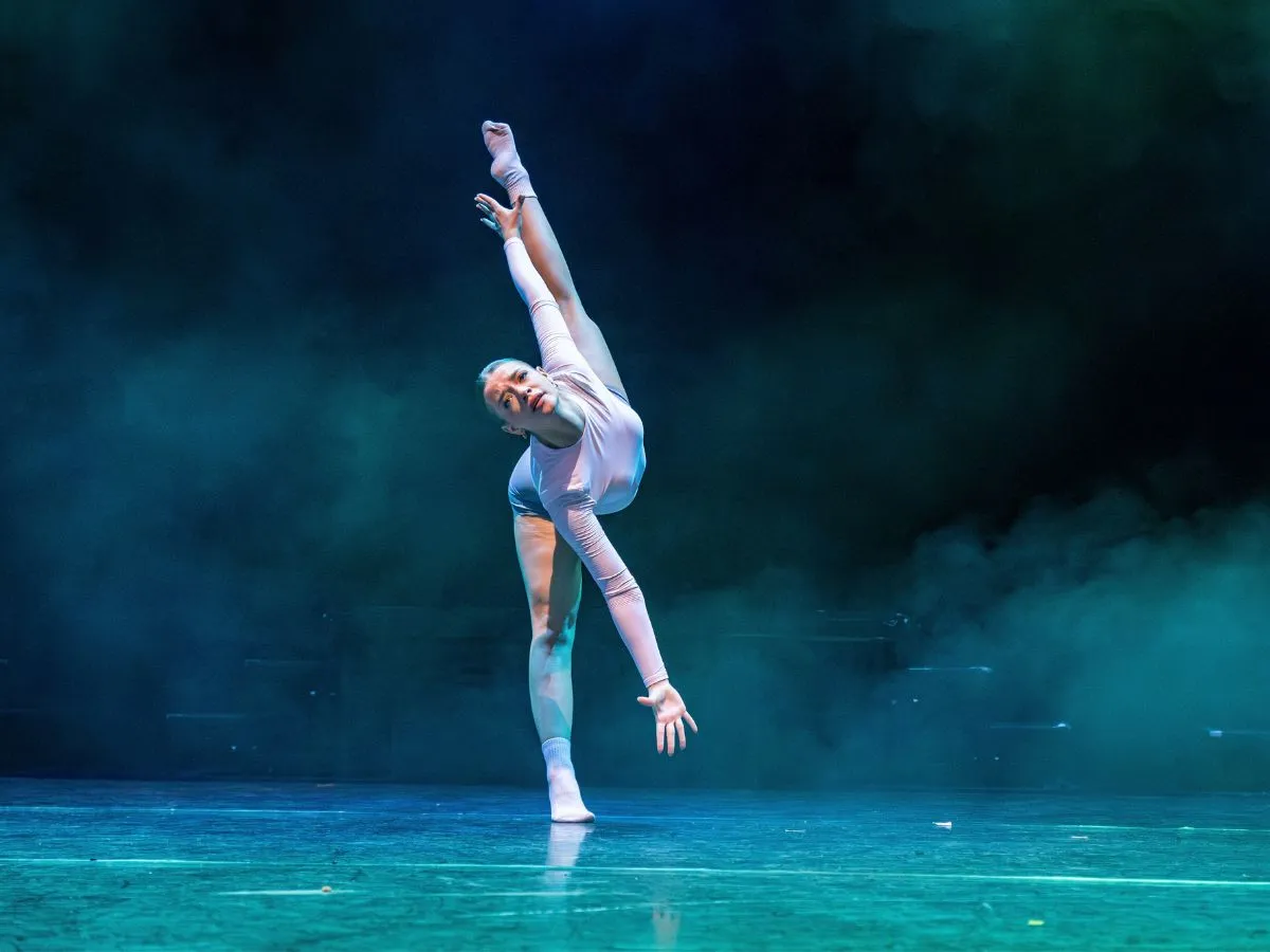 A dancer on stage in a deep tilt extension, one leg raised high, lit in teal haze.