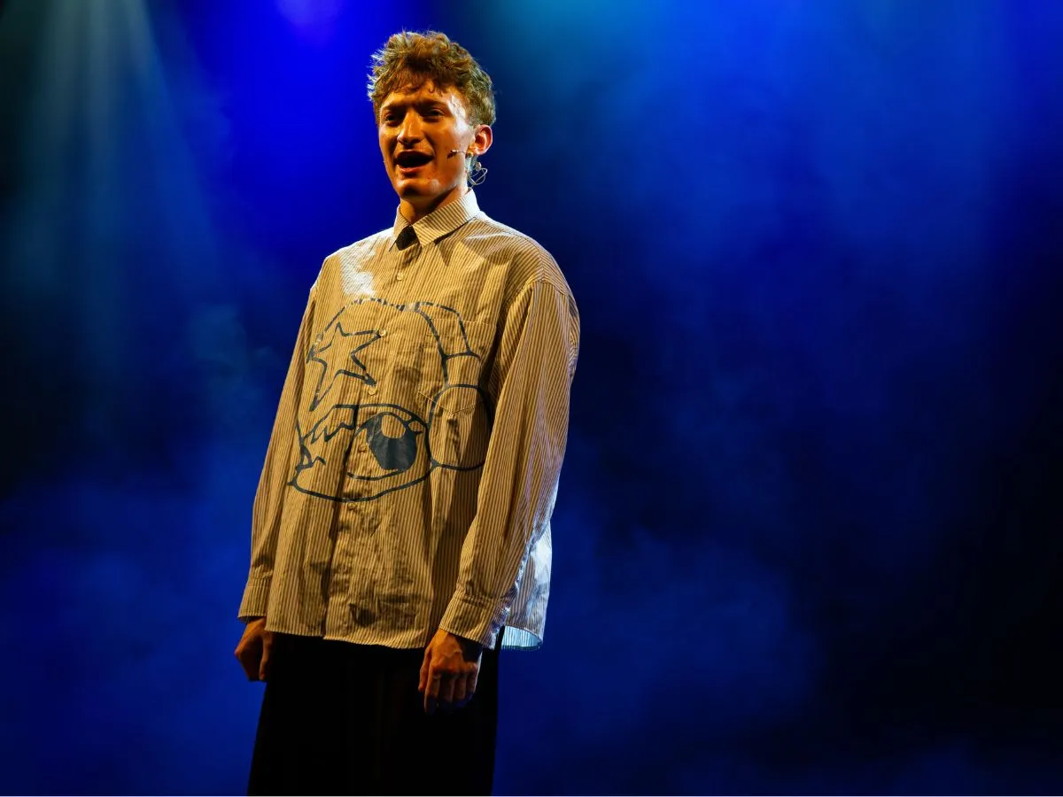 A performer in a striped shirt sings on stage under blue light, wearing a headset microphone.