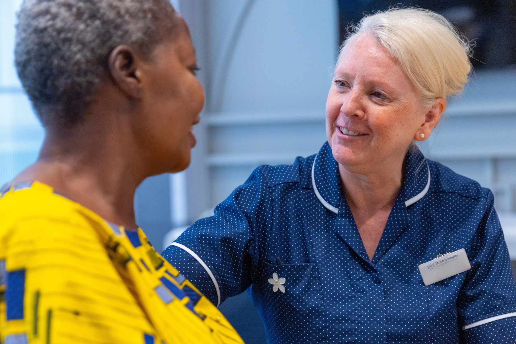 Wellbeing Centre Manager Julie sits across from a patient as they smile and chat. Julie is wearing a blue burses uniform which has little polka dots on. The patient is wearing a yellow dress.