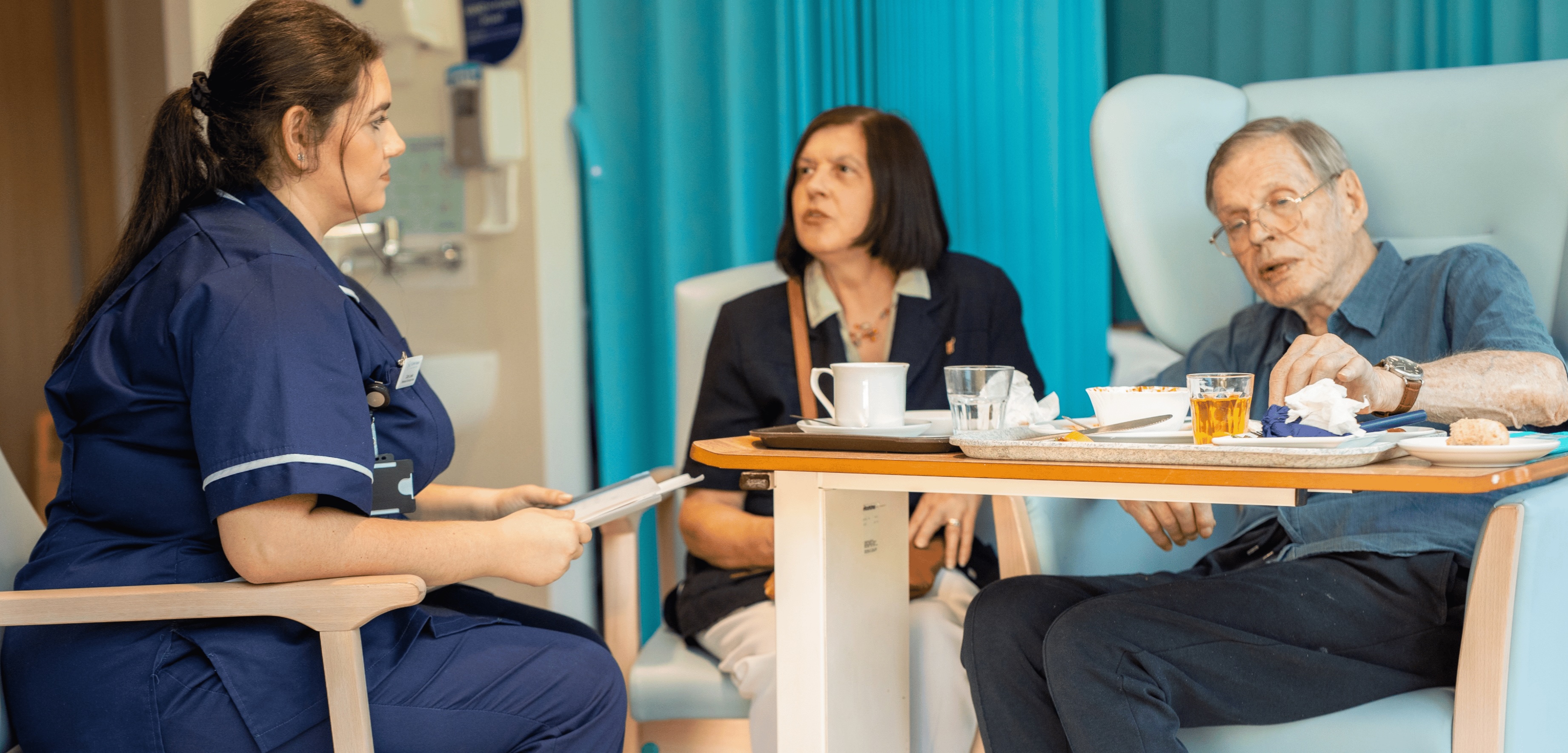Nurse speaking with an elderly man and a woman seated around a hospital bedside table
