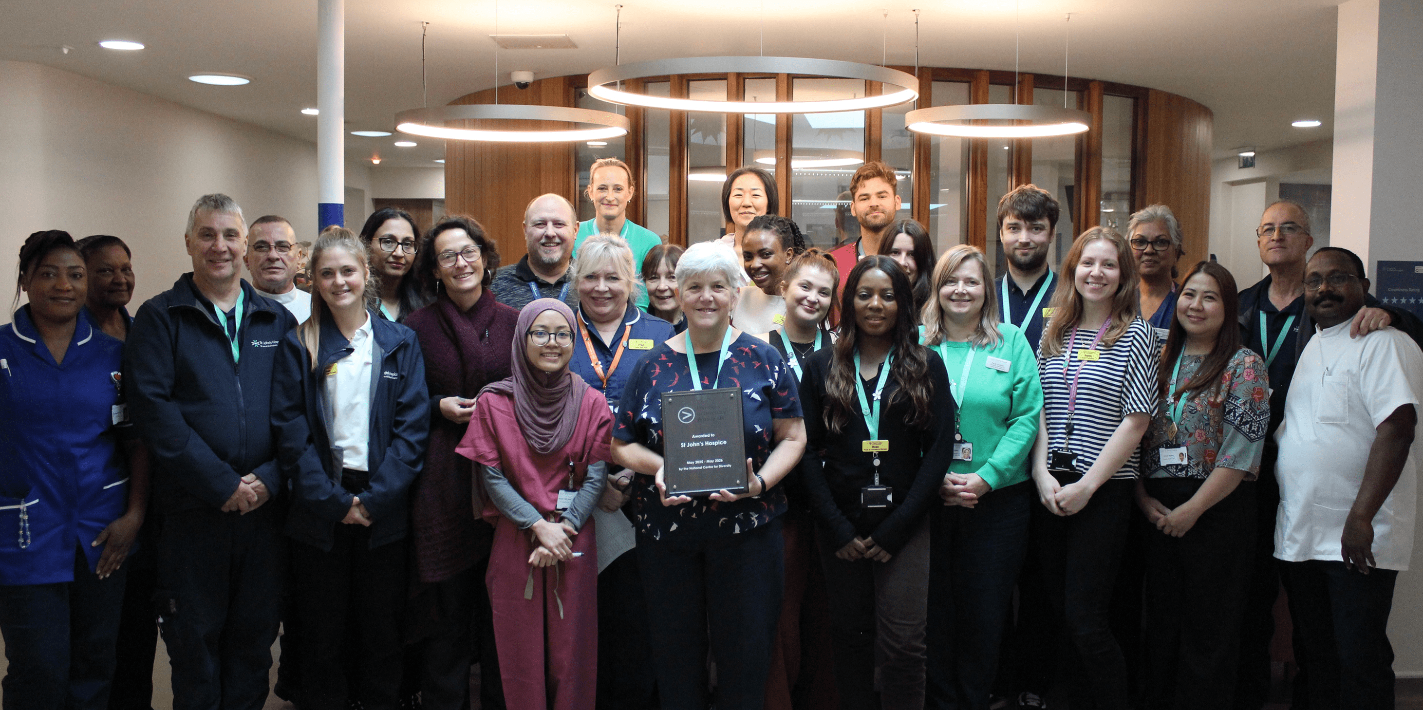 A large group of staff from St John's Hospice huddle together proudly holding the Investors in Diversity Bronze plaque we have been awarded.