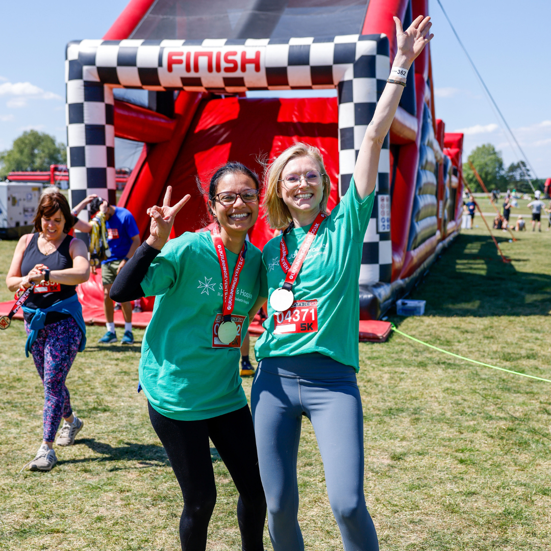 Poppy and her friend pose in front of the giant obstacle course wearing their medals. They are smiling to the camera and doing peace signs.