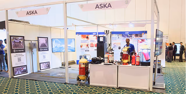 Exhibition booth labeled ASKA displaying fire safety equipment including extinguishers, with a man standing behind the counter.