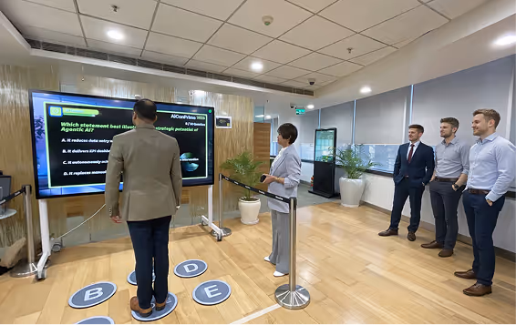 Three men and one woman in business attire participating in an interactive quiz with large screen and floor markers labeled B, C, D, and E in a modern office space.