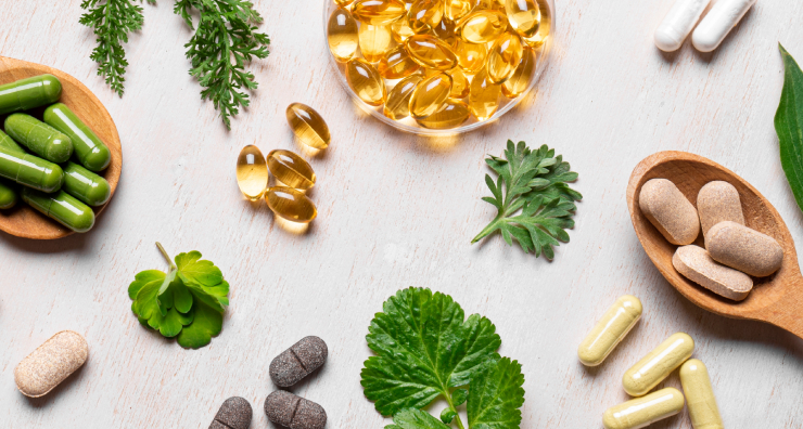 Assorted vitamins and supplement capsules arranged on wooden spoons with fresh herbs on a light wood surface.