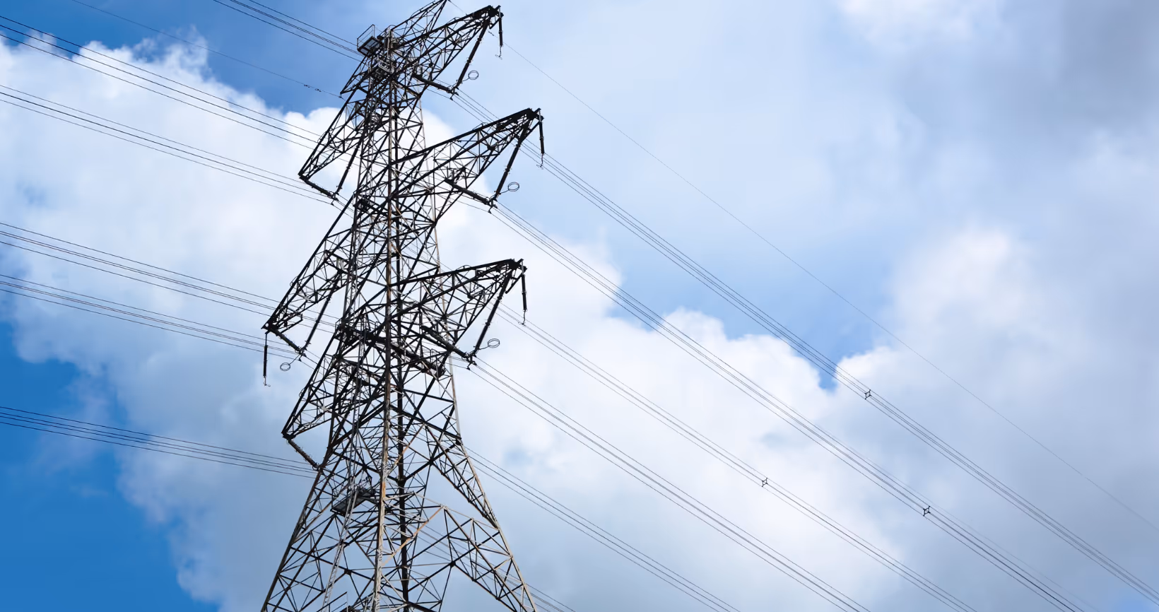 Upward angle photograph of tall electrical transmission tower structure against blue sky with white clouds, showing multiple levels of power line connections and lattice framework construction