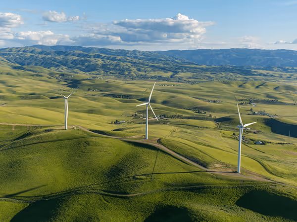 Aerial photograph of wind farm installation with three large white wind turbines on rolling green hills with mountains in background and scattered buildings in rural landscape