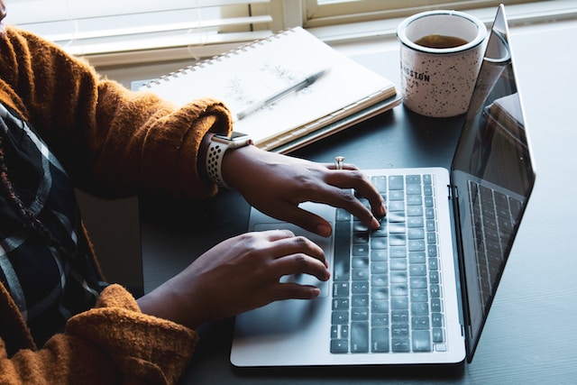 A photo of a person in a dark yellow sweater typing on a laptop that’s placed on a table next to a mug