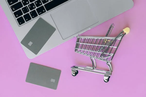 A small silver shopping cart next to a laptop with silver credit cards placed on top