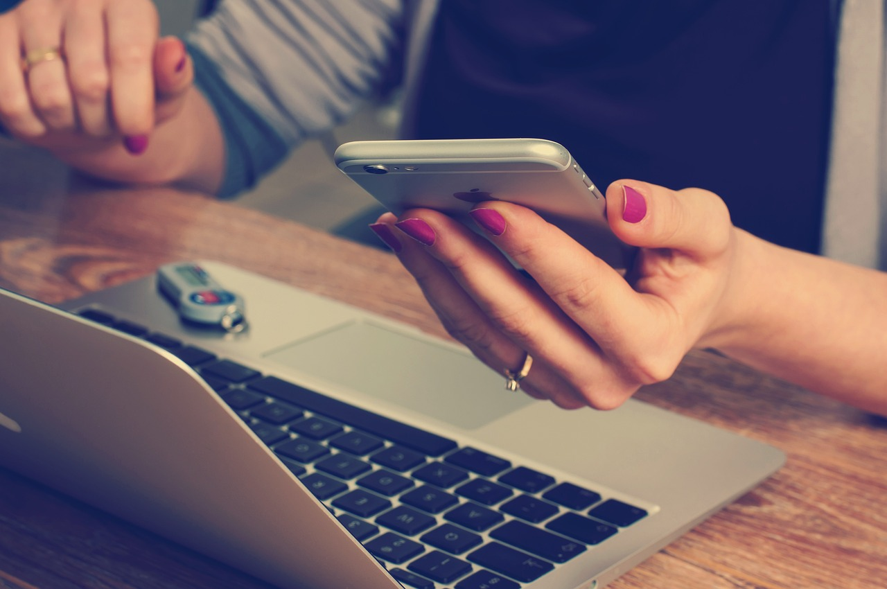 A close-up photo of a woman holding an iPhone in her hand and a Macbook placed on a wooden table