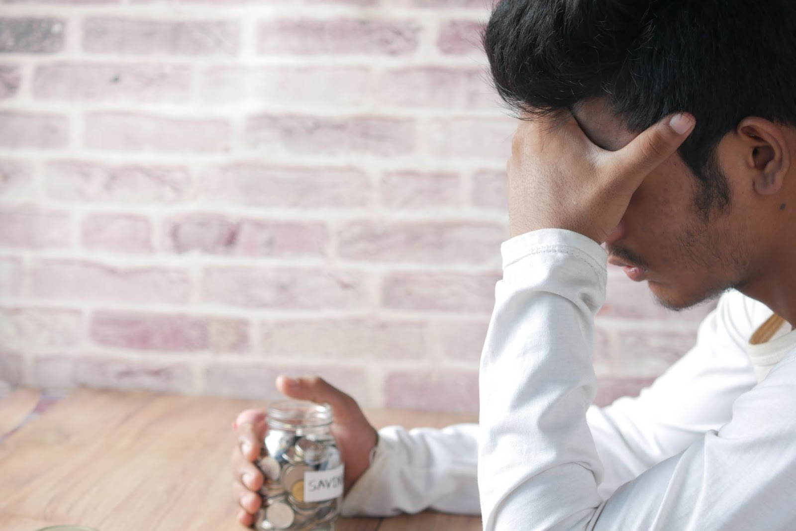 A photo of a person holding their head with one hand and a jar of coins with the other