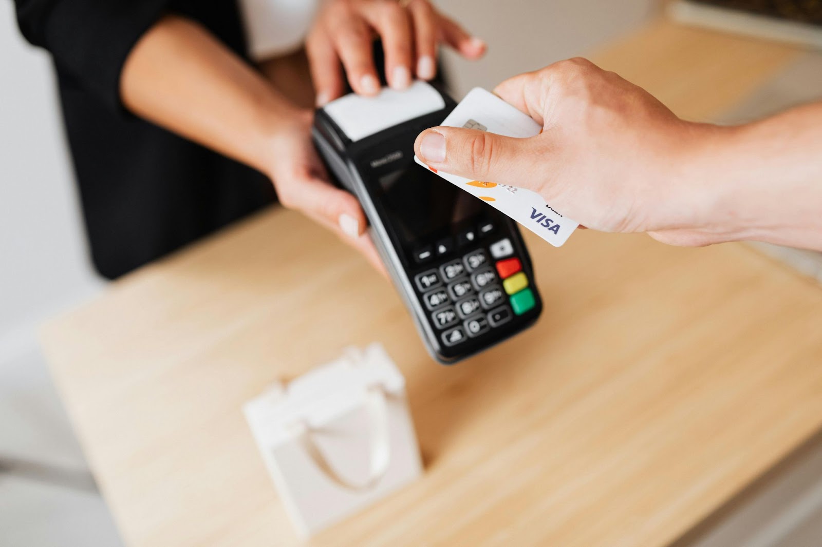 A photo of a hand holding a Visa credit card near a card reader for contactless payment, with a small shopping bag on the counter