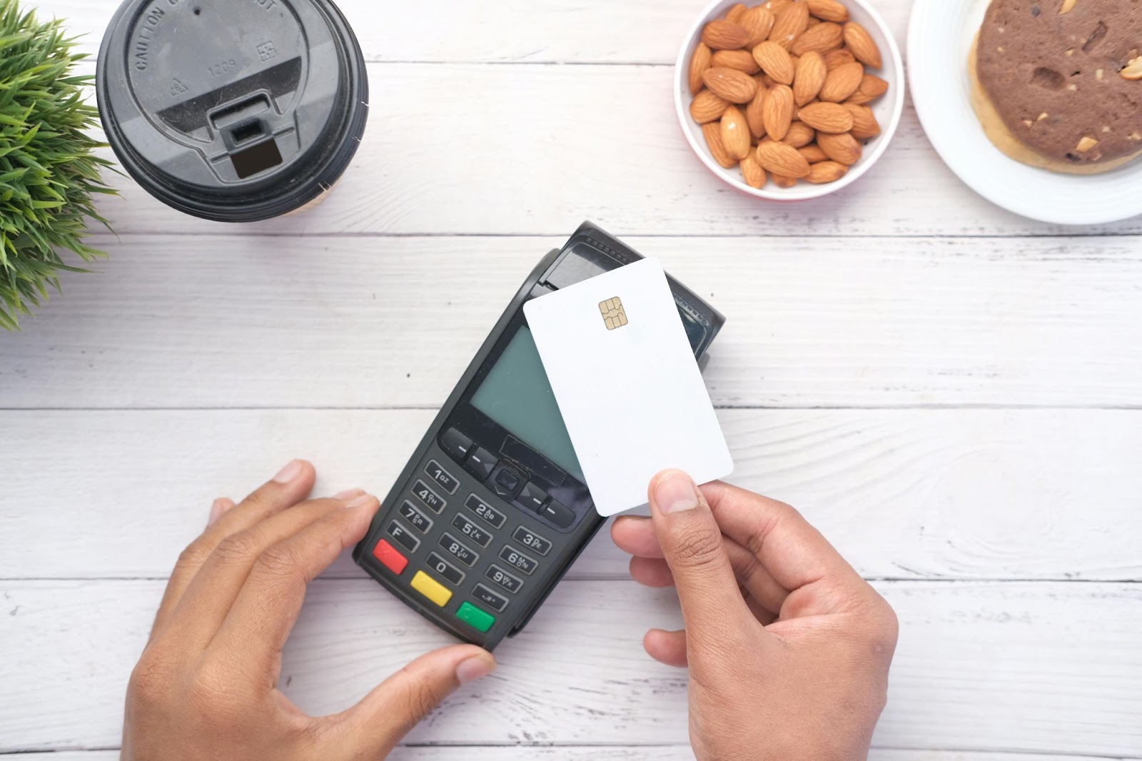 A person's hands holding a white credit card over a payment terminal, with almonds, a cookie, and a coffee cup lid nearby