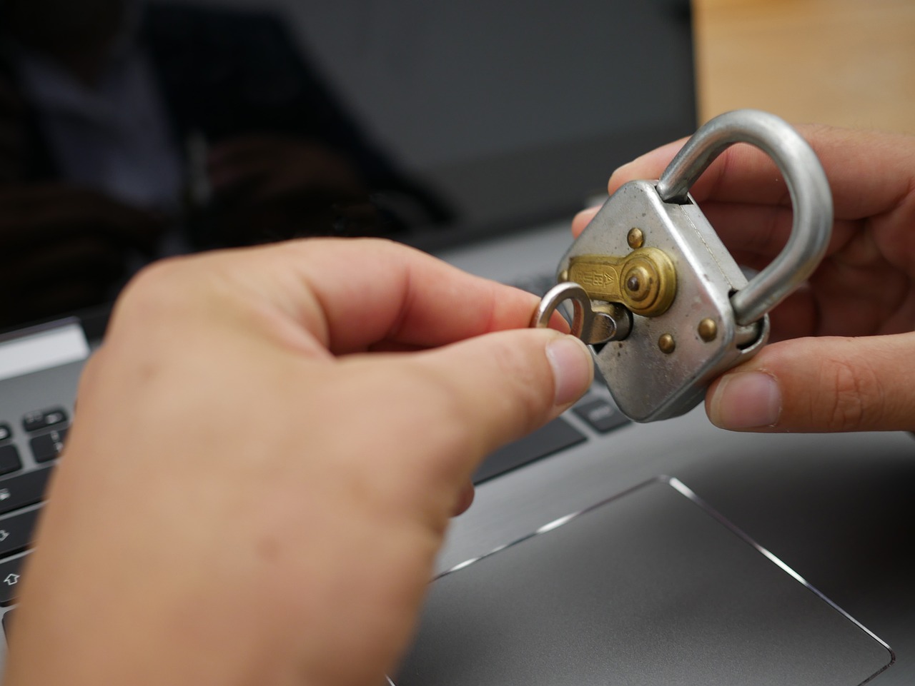 An image of a person's hands trying to open a padlock, with an open laptop in the background