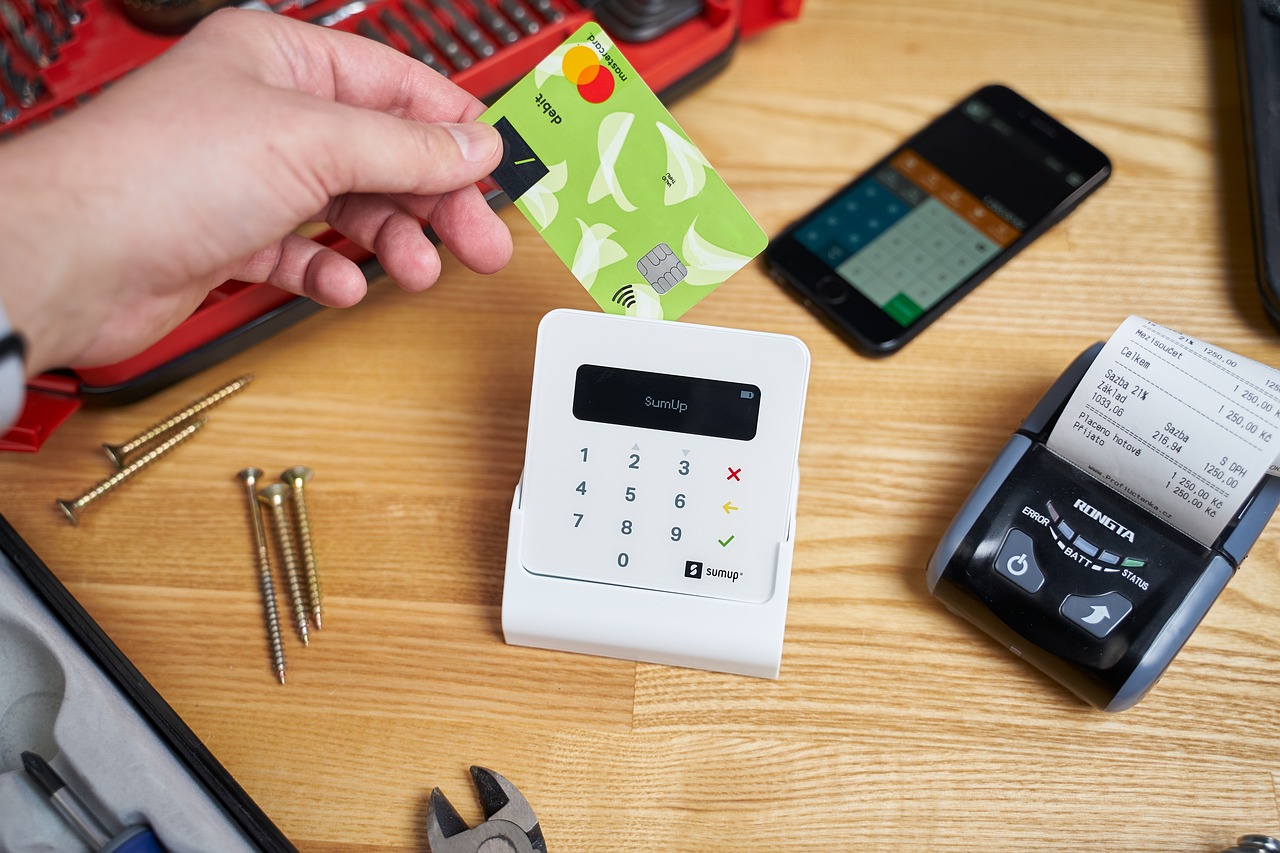 A hand holding a payment card over a card reader, with a receipt printer and a smartphone showing a calculator on the table