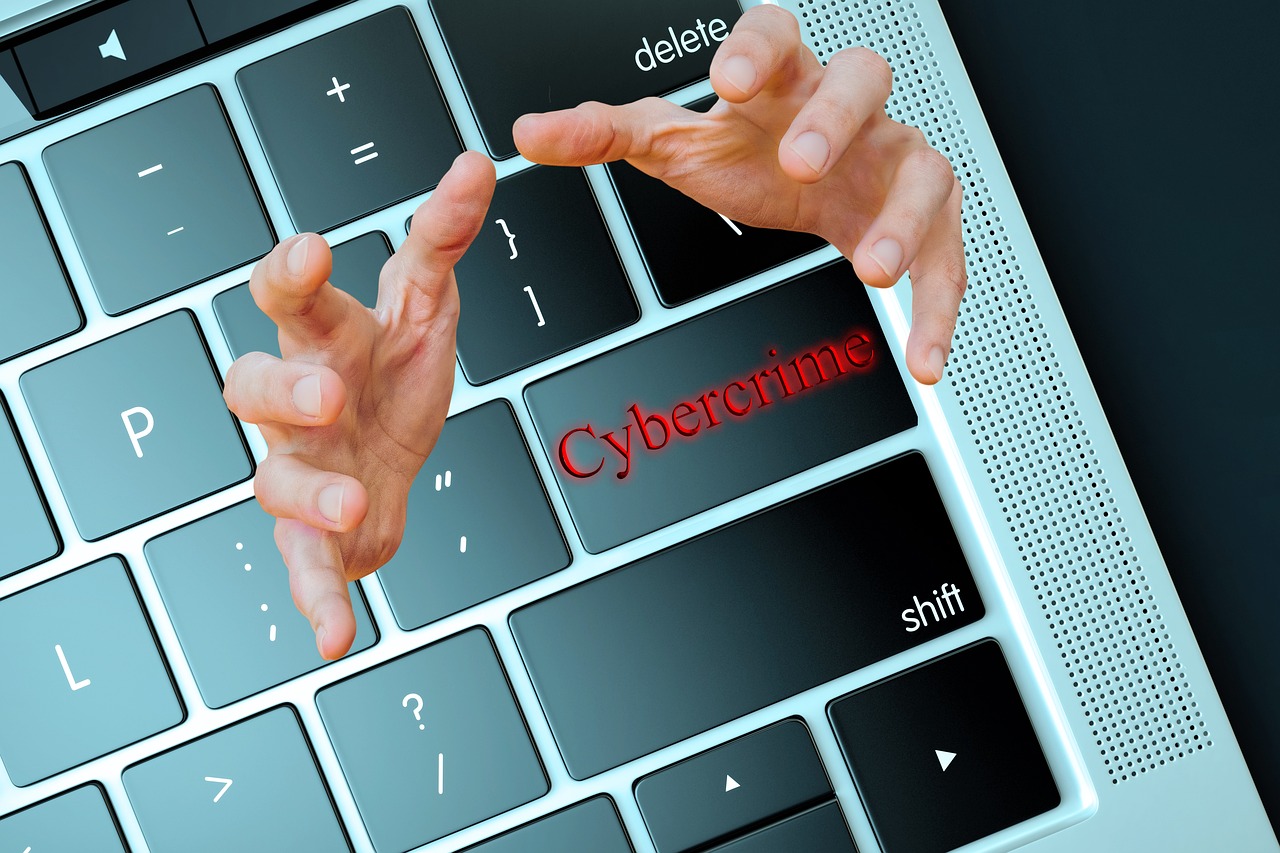 A laptop keyboard with the word "Cybercrime" highlighted in red on one key, and hands reaching out toward it