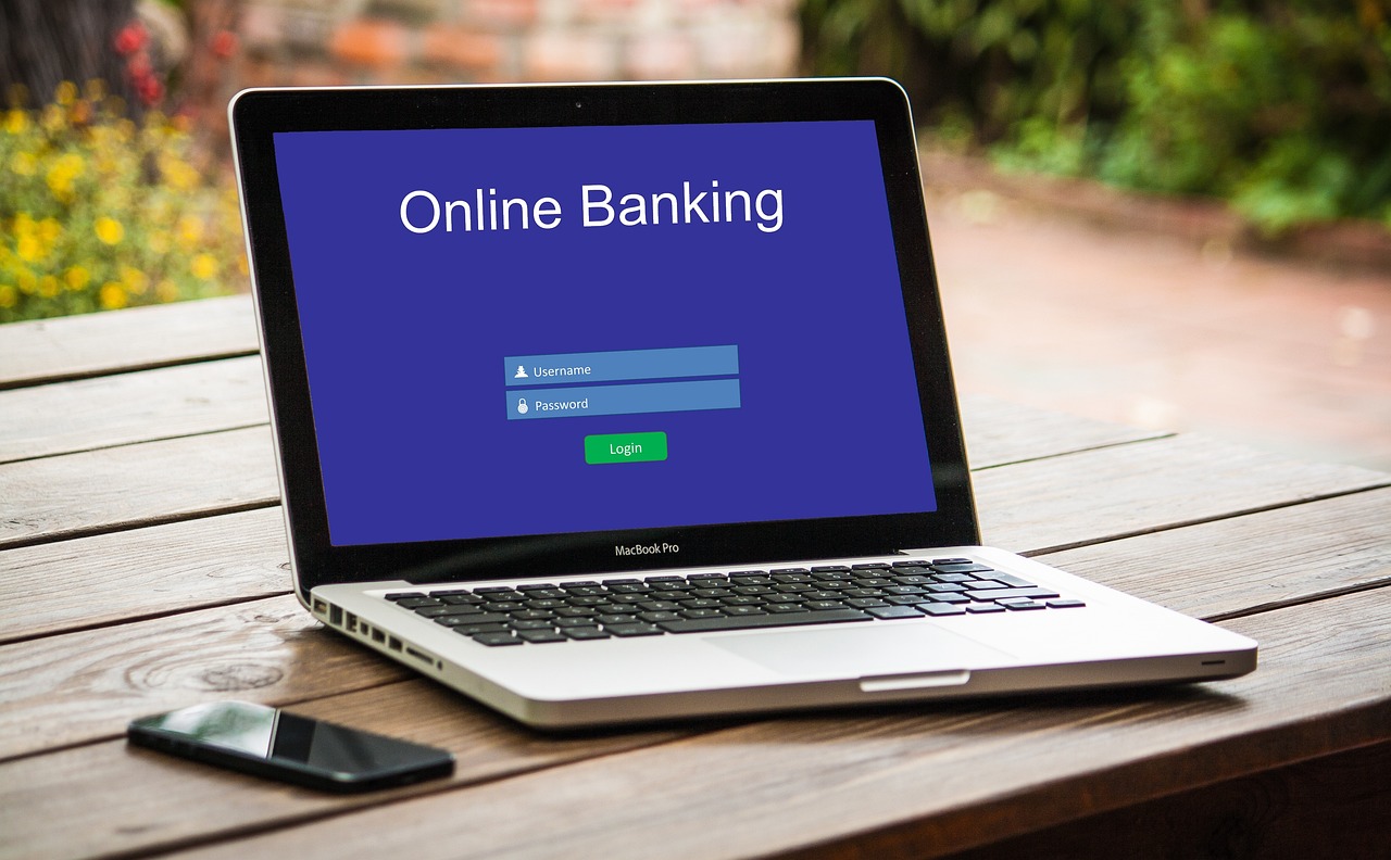 A photo of an open laptop on a wooden table, displaying an online banking login page, with a smartphone beside it