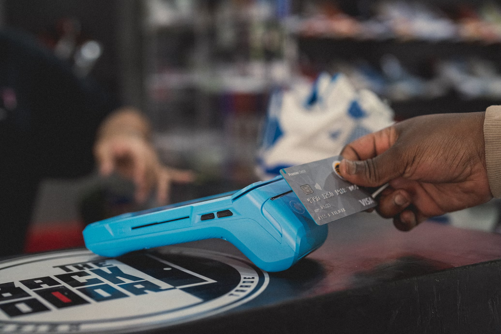 A cropped photo of a hand holding a payment card to a blue point-of-sale terminal placed on a countertop