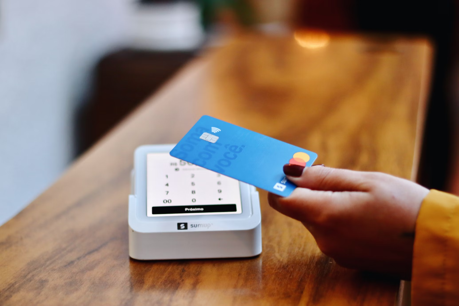 A cropped photo of a person holding a blue payment card over a white card reader placed on a wooden surface