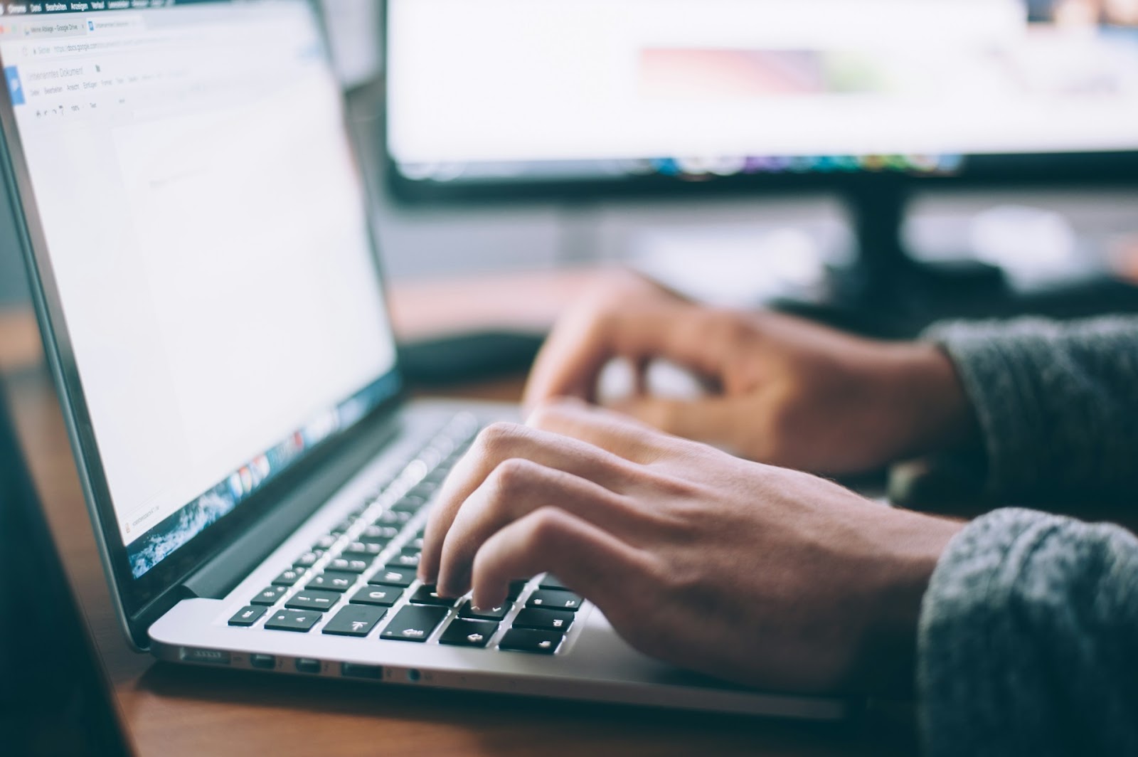 An image of a person typing on a laptop with a computer monitor visible in the background