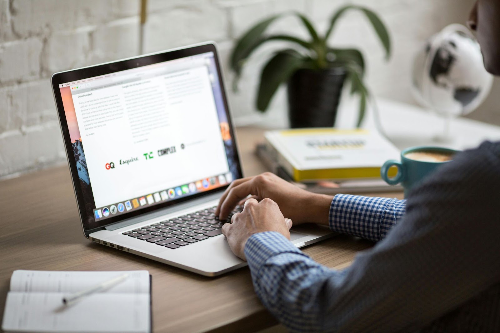 An image showing a person working on a laptop placed on a wooden table with a notebook and a cup of coffee next to it