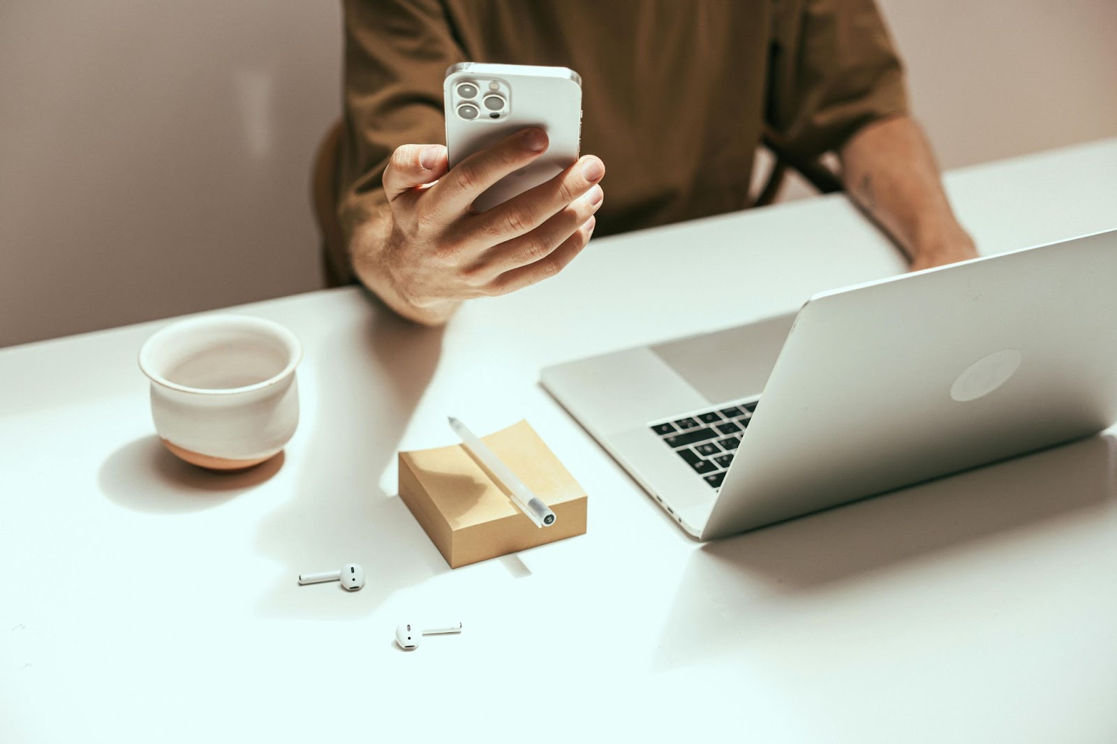 A close-up photo of a person sitting at a white desk and holding a smartphone in one hand while using a laptop