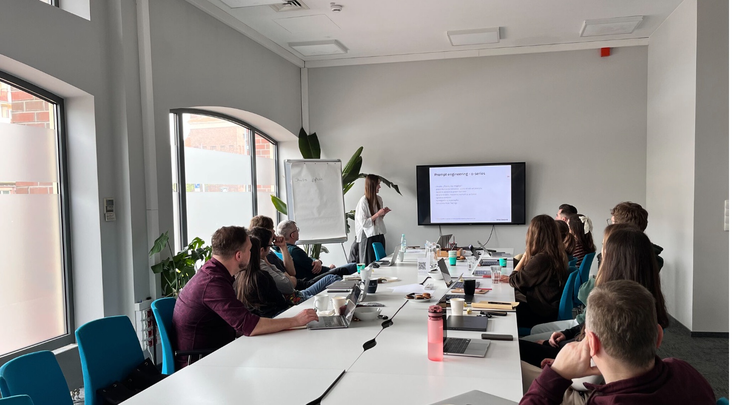 A group of people seated around a long conference table in a bright meeting room, watching a presenter at the front who is pointing to a slide displayed on a wall-mounted screen.