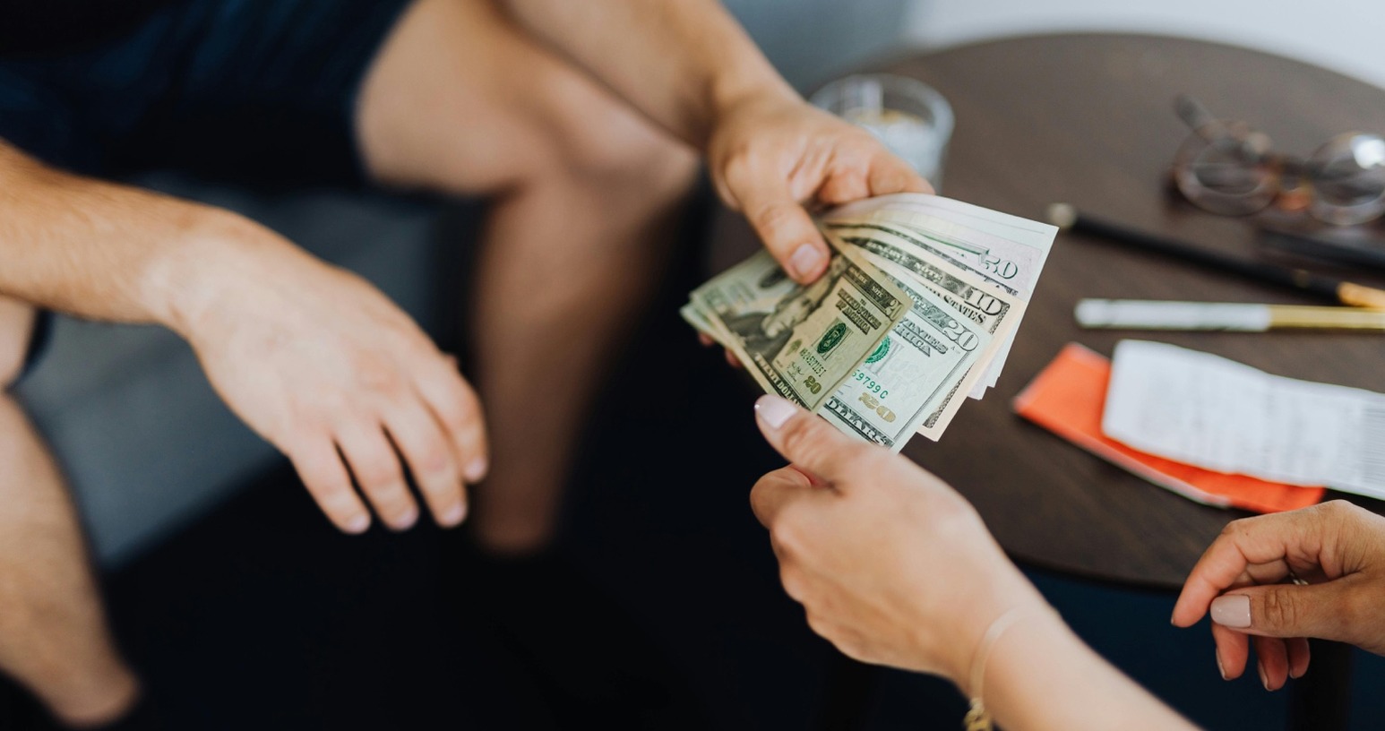 Two people seated at a table, one handing a stack of U.S. dollar bills to the other. A receipt and wallet lie on the table beside them.