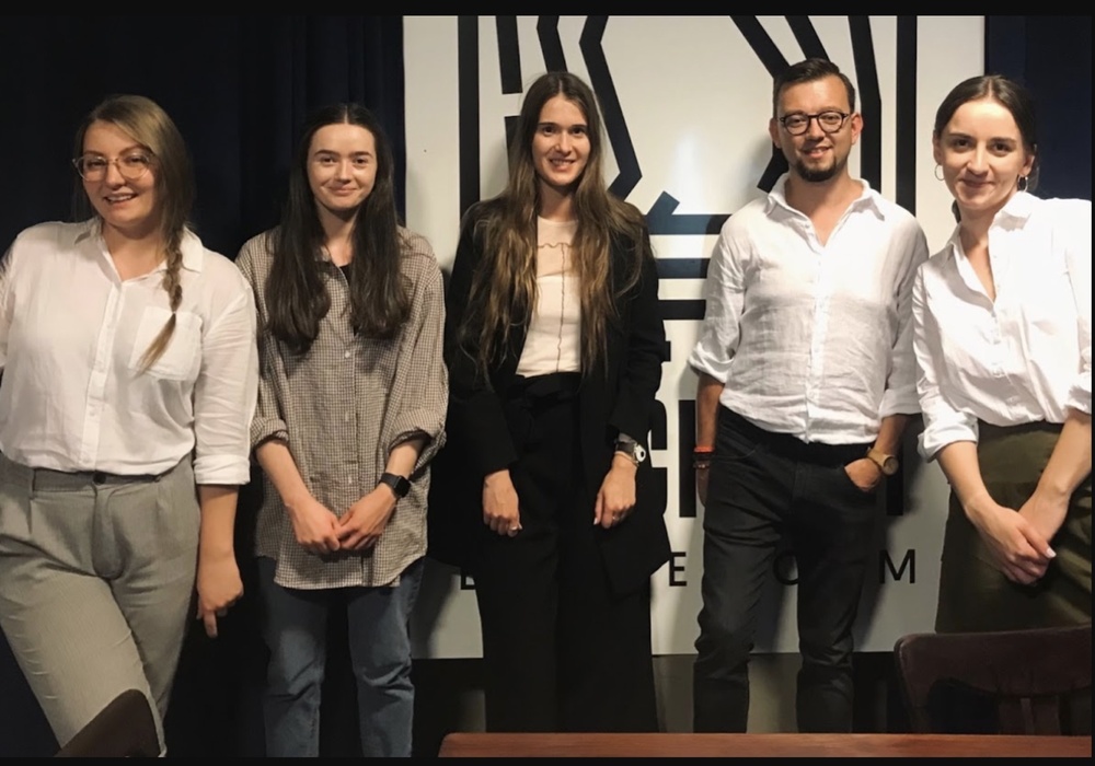A photograph of five young people—four women and one man—standing side-by-side on a stage against a dark background with a graphic pattern visible behind them. They are dressed in smart-casual attire, smiling at the camera, likely after an event or presentation.