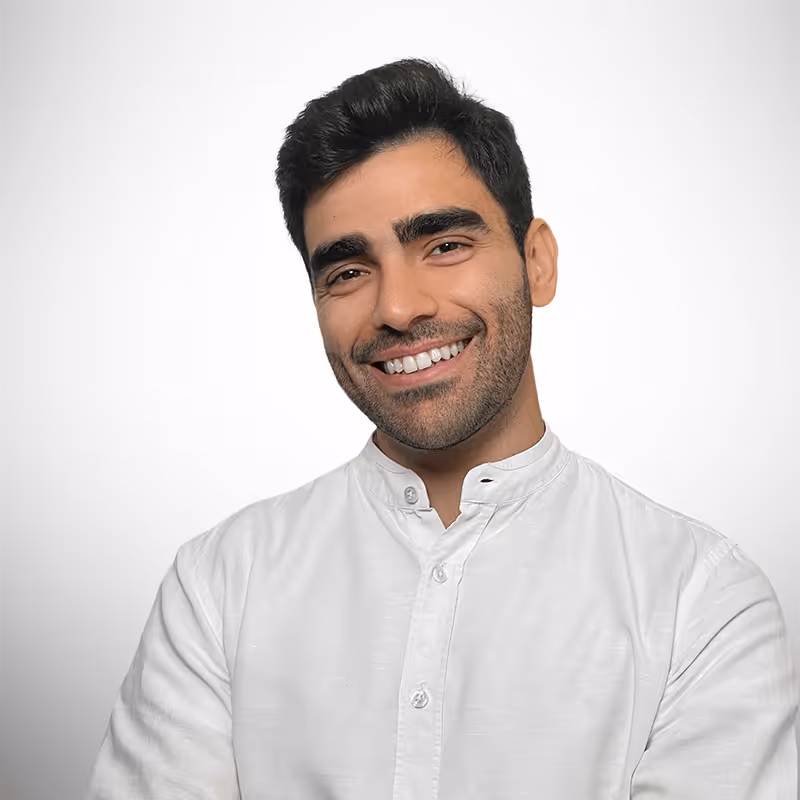 A young man with short black hair and a well-groomed beard smiles warmly at the camera. He is wearing a white mandarin-collar shirt with the top button undone. His arms are crossed, and he has a relaxed and confident posture. The background is a neutral gradient.