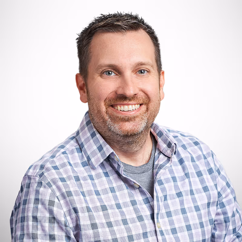 A man with short, slightly spiked brown hair and light stubble smiles warmly at the camera. He is wearing a plaid button-up shirt in shades of purple, white, and gray, layered over a gray t-shirt. His posture is relaxed and approachable, with a neutral gradient background.